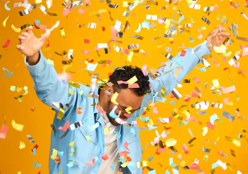A person in a light blue jacket raises their arms in celebration as colorful confetti falls on a bright yellow background.