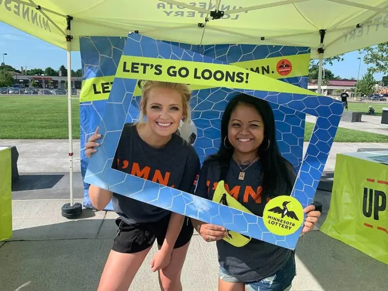 Two people smile, holding a blue "Let's Go Loons!" frame under a Minnesota Lottery-branded yellow tent in a grassy park setting.