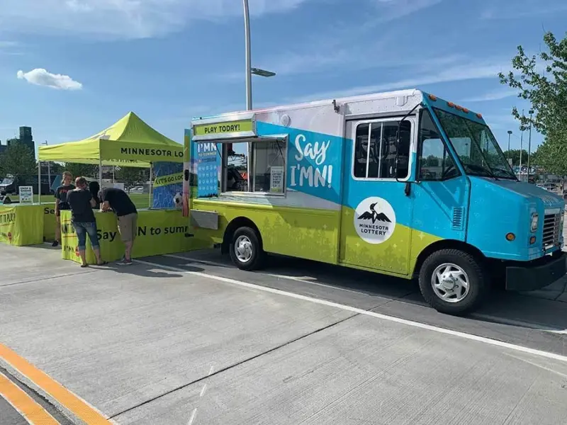A colorful "Say I’MN!" food truck and a yellow Minnesota Lottery tent are set up outdoors, with people gathering nearby.
