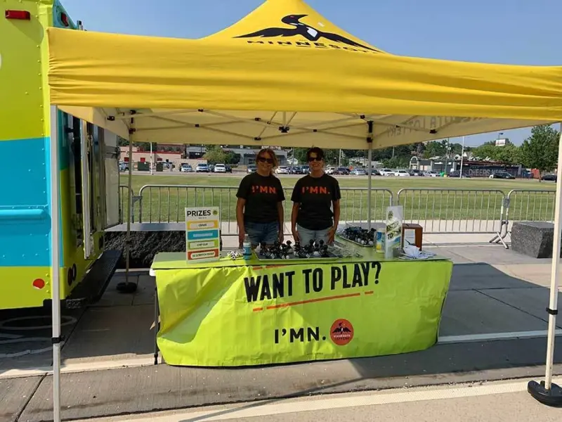 Two people under a yellow tent with a "WANT TO PLAY? IM.N" banner; table displaying items and prize list, blue-green trailer in background.