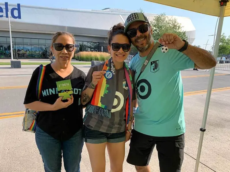 Three people in sports gear, posing by a stadium. The middle person holds a medal.