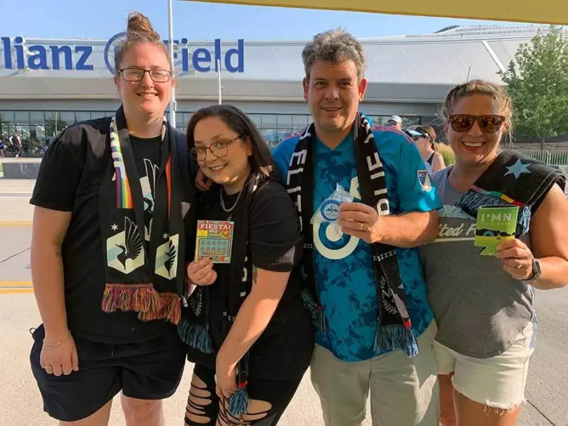 Four people wearing soccer scarves smile in front of Allianz Field, holding small items. The stadium is visible under a sunny sky.