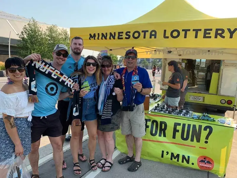 Six people smile in front of a yellow Minnesota Lottery tent, holding tickets and wearing soccer scarves, with a table of promotional items nearby.