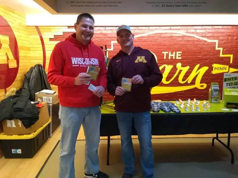 Two smiling men hold packets, standing by a table in a yellow and maroon room with "Barn" signage. One sports a red Wisconsin hoodie, the other Minnesota gear.