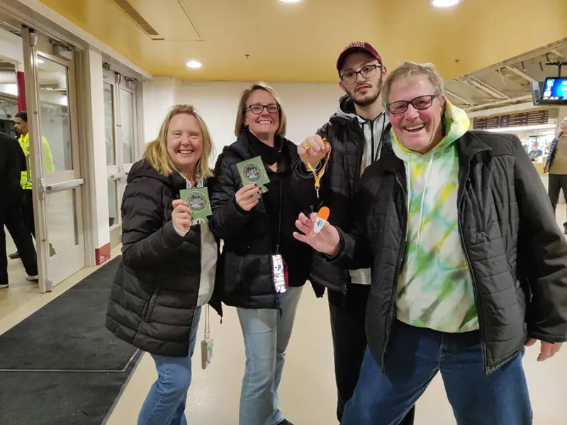 Four smiling people indoors; two hold green passes, one holds a lottery ticket and keychain. They are warmly dressed and appear happy.