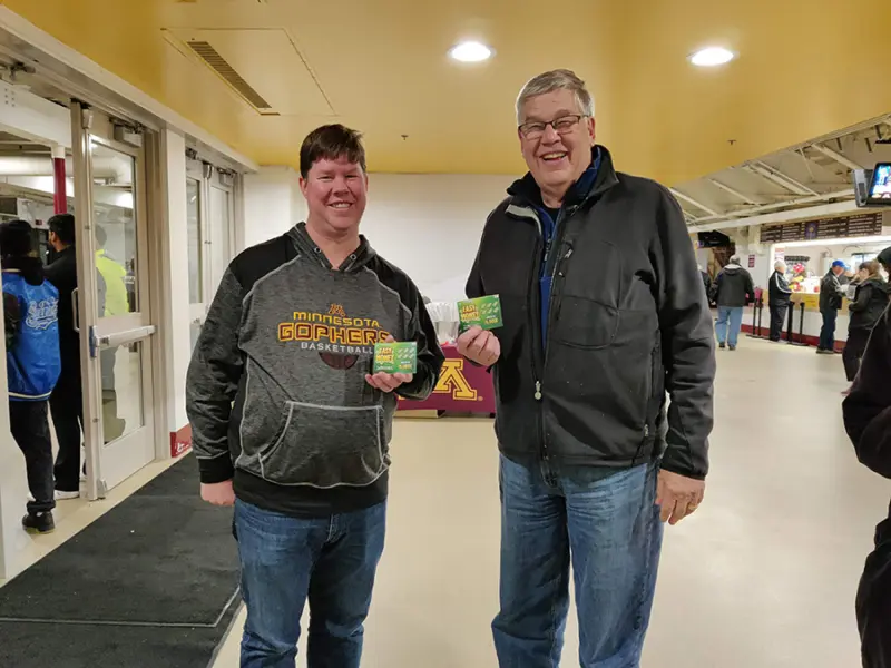 Two people indoors at a sports venue smiling and holding Minnesota Lottery tickets. One wears a Gophers hoodie; others are nearby, near entrance doors.