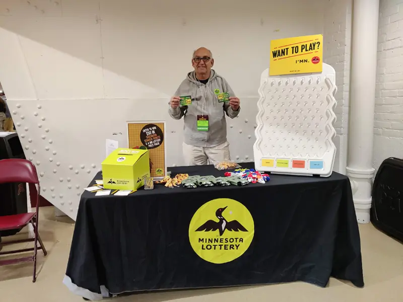 Person holds Minnesota Lottery tickets behind a branded table with Scratch Games, Lotto, Print-N-Play, Raffle tickets. "Want to Play?" sign above.