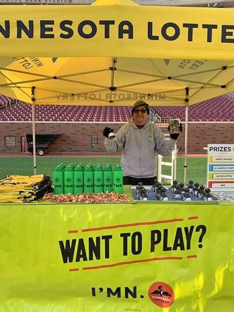 A person smiles under a yellow Minnesota Lottery tent, holding tickets. A table displays green water bottles, promo items, and a "Want to Play? I'm in" sign.