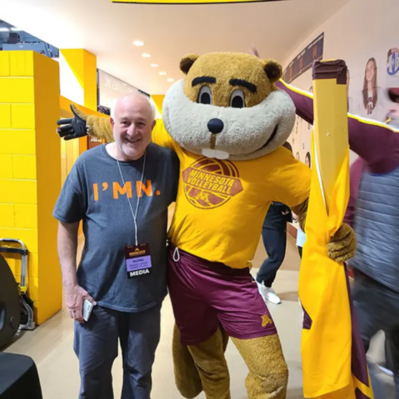 A smiling person with a media pass stands indoors beside a mascot in a Minnesota volleyball shirt, holding a yellow flag, promoting lottery games.