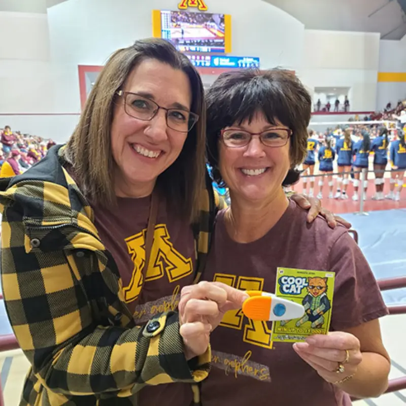 Two women with glasses, wearing Minnesota shirts, smile in a sports arena holding a lottery ticket with a cartoon and puppet. Volleyball players on court behind.