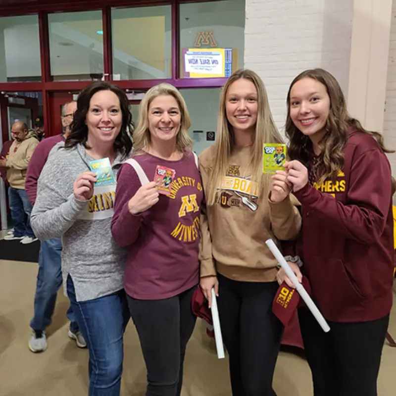 Four women in maroon and gold hold sports tickets indoors, smiling excitedly. The background features a "M Bank" sign.