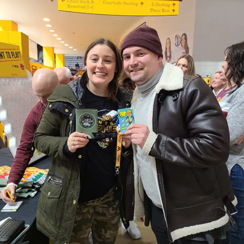 A smiling woman in a green jacket and a man in a brown jacket with maroon beanie hold badges at an indoor event, showcasing Minnesota Lottery excitement.