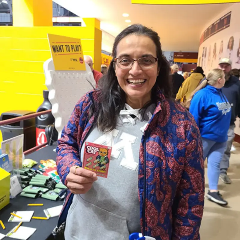 A smiling person holding a comic-style Minnesota Lottery card in a busy indoor area, wearing glasses and patterned jacket over a hoodie.