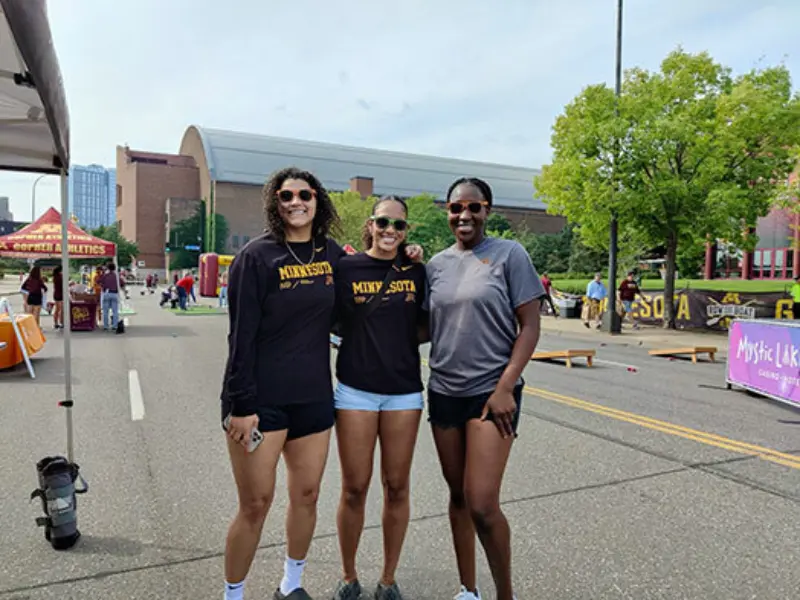 Three women in casual athletic wear, two with "Minnesota" shirts, smile at an outdoor event with booths; promoting Minnesota State Lottery excitement.
