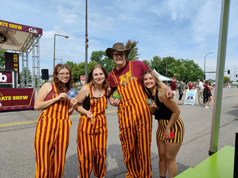 Four smiling people at a fair, in matching striped outfits. Behind them, a stage promotes Minnesota State Lottery games: Scratch, Lotto, Raffle, 2nd Chance.