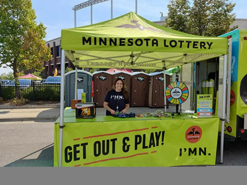 Person at Minnesota Lottery booth with "GET OUT & PLAY! I'M IN" sign, prize wheel, promo items. Portable toilets and trees in the background.