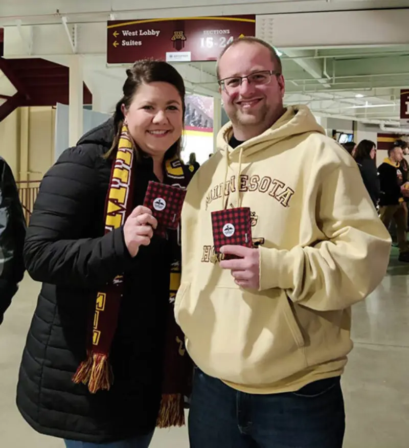 Two people indoors, smiling. Left: dark coat and scarf; Right: Minnesota hoodie. Both hold items featuring lottery logos in a venue with signs above.