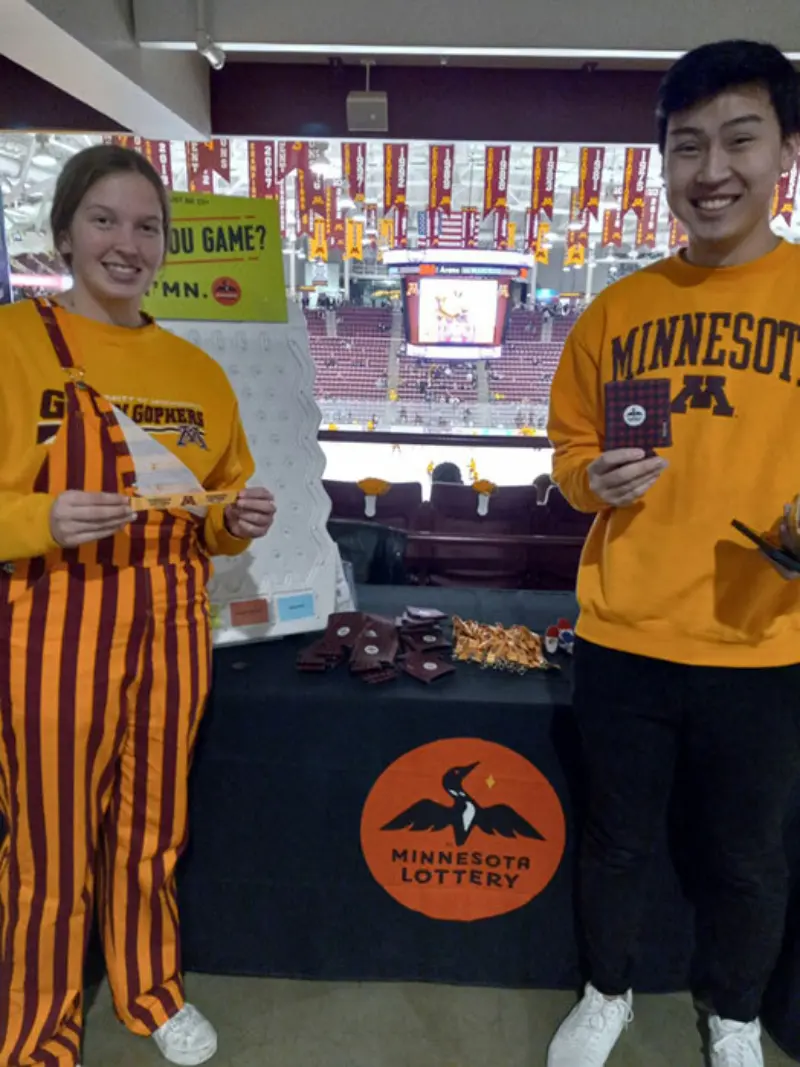 Two University of Minnesota fans stand by a Minnesota Lottery booth at a sports venue, showcasing lottery tickets. Hockey rink and screen in the background.