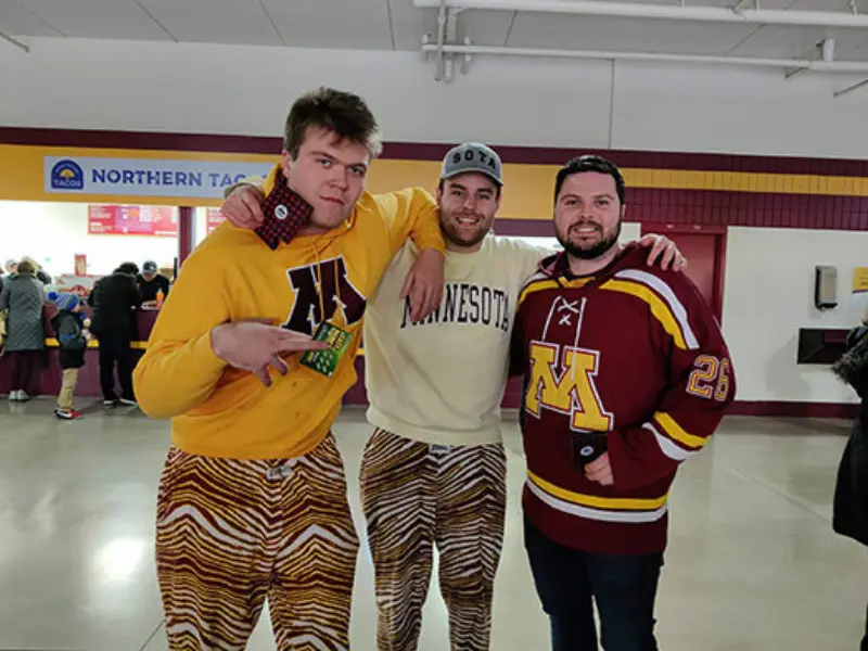 Three people at a sports venue; two in Minnesota-themed outfits. One wears a SOTA hat. A concession stand is visible in the background.