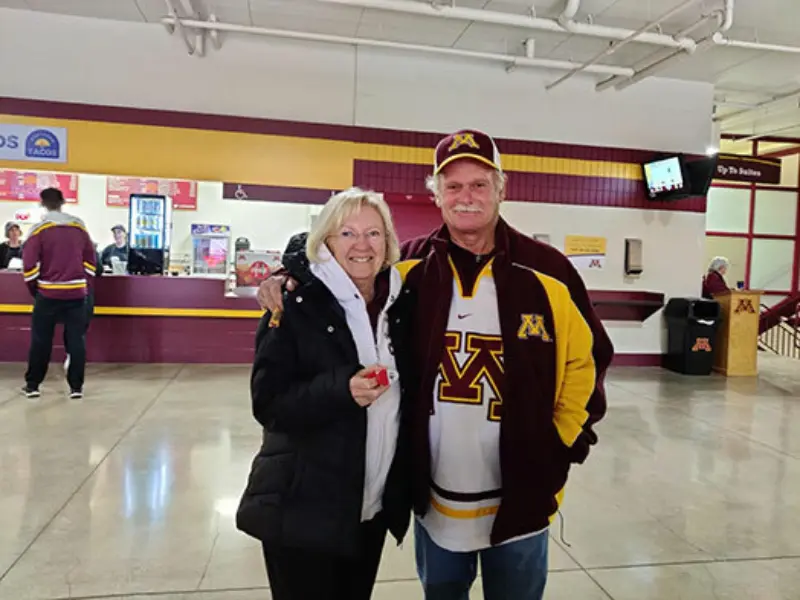 Man in Minnesota Gophers gear with woman at sports venue. Concession stand visible. They're excited about MN Lottery: Scratch, Lotto, Print-N-Play & more.