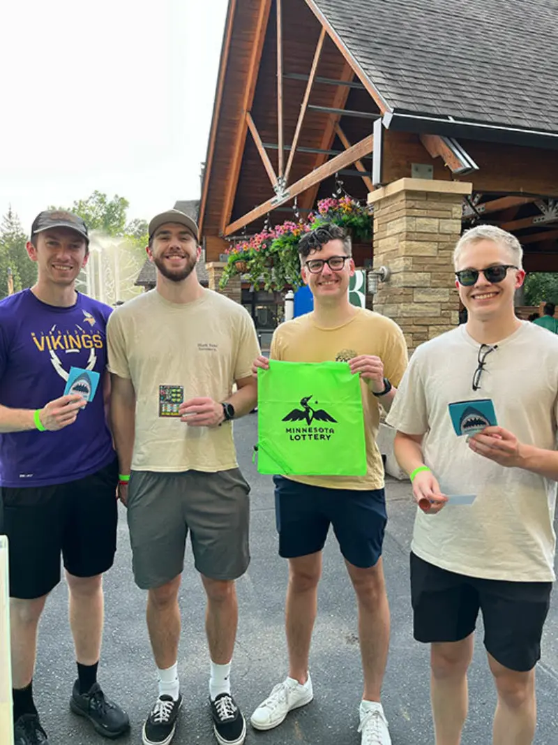 Four men smile outside a wooden building; one with a green Minnesota Lottery bag, others hold lottery tickets, enjoying a sunny day.