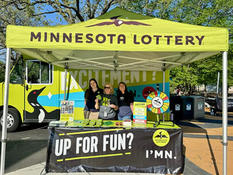 Three people at a Minnesota Lottery booth with a wheel, promotional items, and loon banner, set outdoors with trees and a lottery truck in the backdrop.