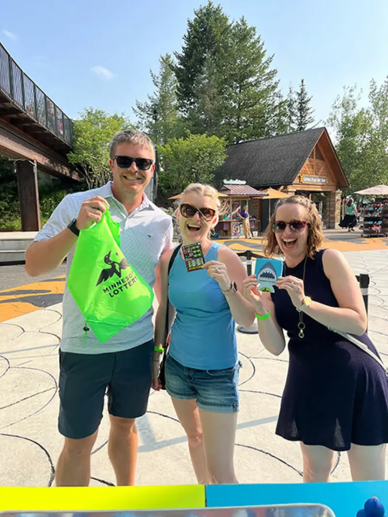 Three people outdoors: one with a green bag, another with a scratch-off ticket, and the third with a blue keychain; trees and building behind.