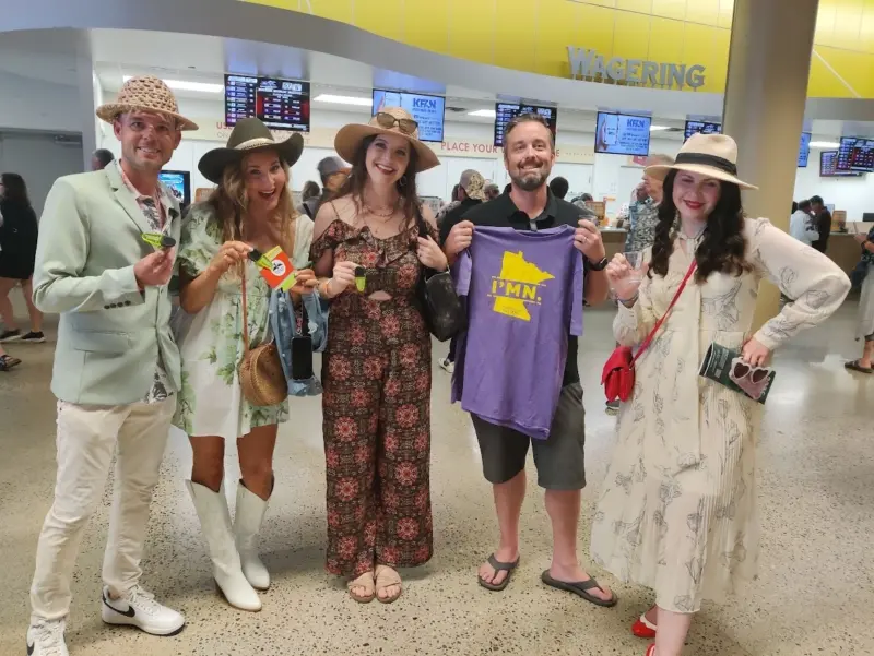 Five smiling people at an indoor racetrack, one holding a purple Minnesota shirt; betting boards and kiosks in the background.