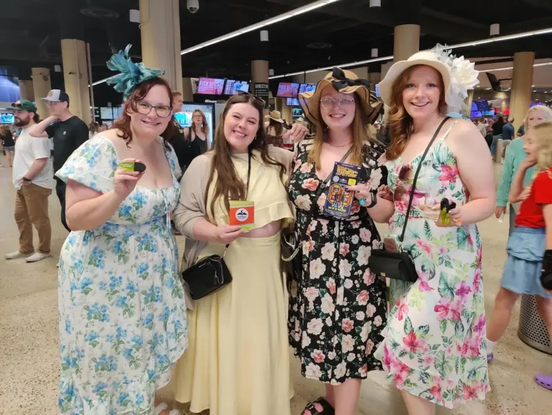 Four women in floral and pastel dresses with hats smile indoors, holding items, with people and screens in the busy background.