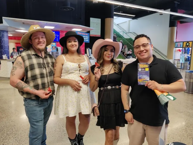 Four people in Western outfits pose indoors, smiling. One holds a Minnesota Lottery flyer; others have small red items and an umbrella.