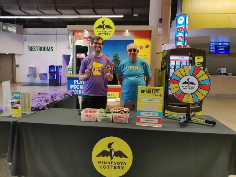 Two people stand at a Minnesota Lottery booth with prize bins, spin wheel, lottery signs, t-shirts, near restrooms and ticket machines.
