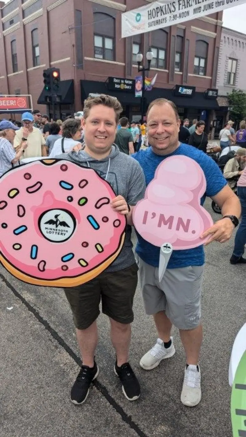 Two smiling men at an outdoor festival hold pink cutouts of a sprinkled donut and “IMN” ice cream scoop; crowds and shops behind them.