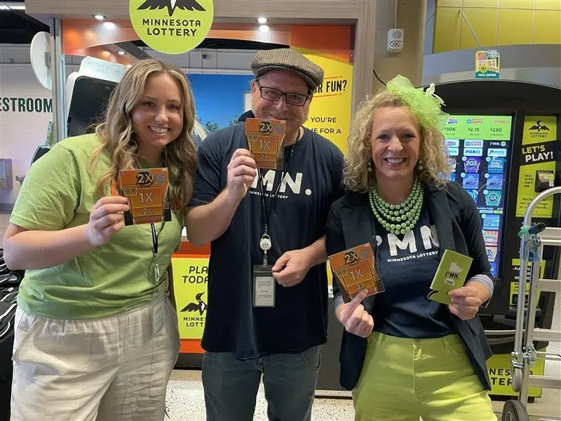 Three people in “MN” shirts display Minnesota Lottery scratch-off tickets in a store, with lottery machines and signs behind them.