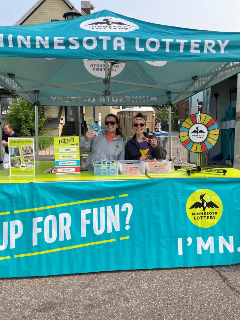 Two people smile under a Minnesota Lottery tent, holding tickets by a prize wheel, promo signs, and giveaway baskets on a blue table.