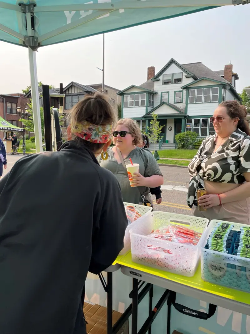 A person serves items at an outdoor Minnesota State Lottery booth while two women chat and hold a drink, houses seen behind them.