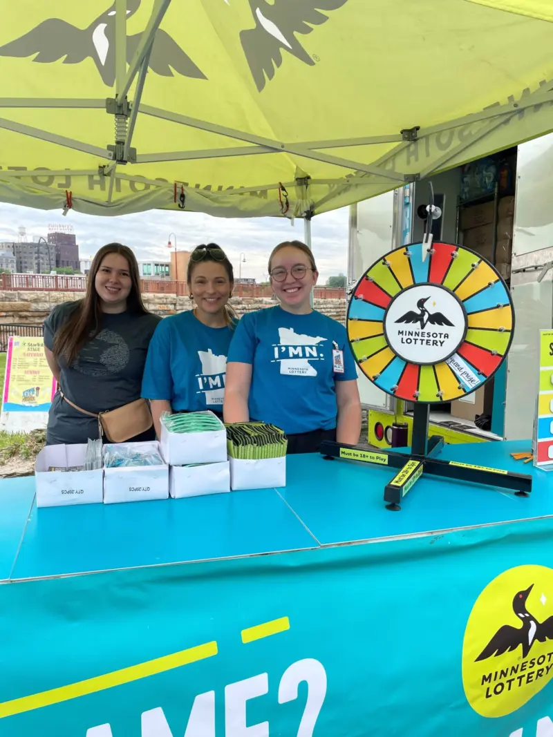 Three people smile behind a Minnesota Lottery booth with a prize wheel, giveaways, and tablecloth under a yellow tent outdoors.