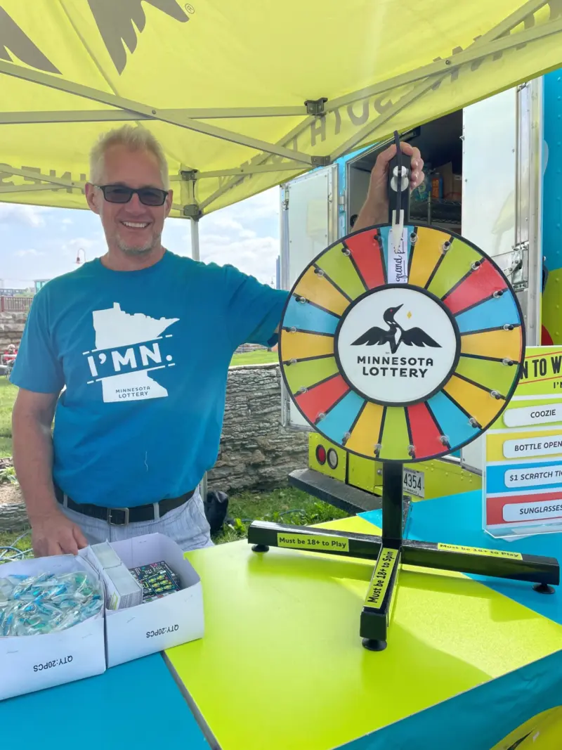Man in sunglasses and blue IMN Minnesota shirt smiles by a colorful Lottery prize wheel under a yellow tent with prize boxes nearby.