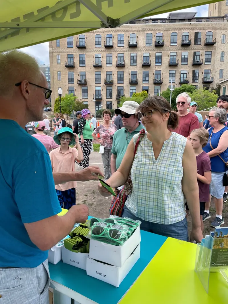 A woman smiles as she gets green sunglasses at a Minnesota Lottery booth, with people in line and apartments in the background.