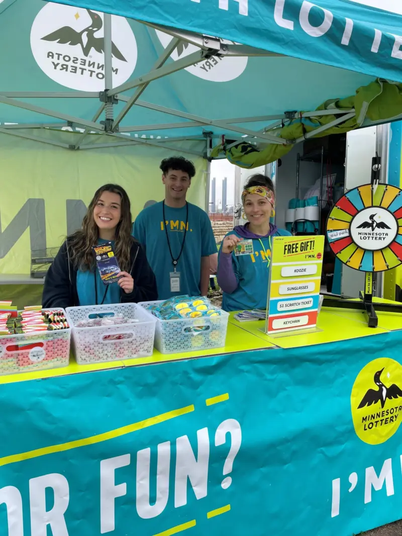 Three people in blue shirts stand at a Minnesota Lottery booth with prize wheel, giveaway baskets, and "I’M IN" banner.