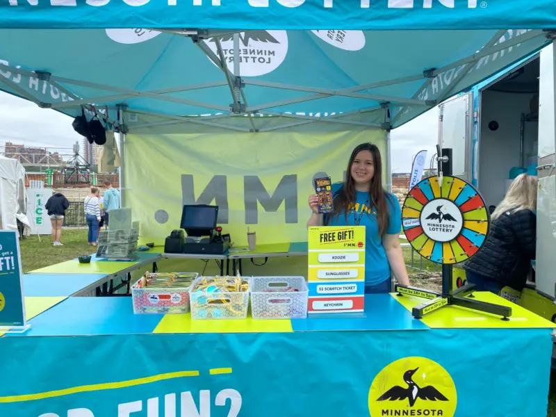 Smiling woman holds a prize by a spinning wheel at an outdoor Minnesota Lottery booth with free gifts and blue-yellow signage.