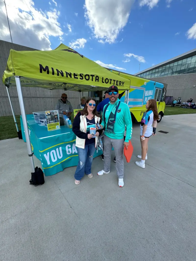 Two people smile in front of a yellow Minnesota Lottery tent at an outdoor event, with a man at the booth and food truck behind them.