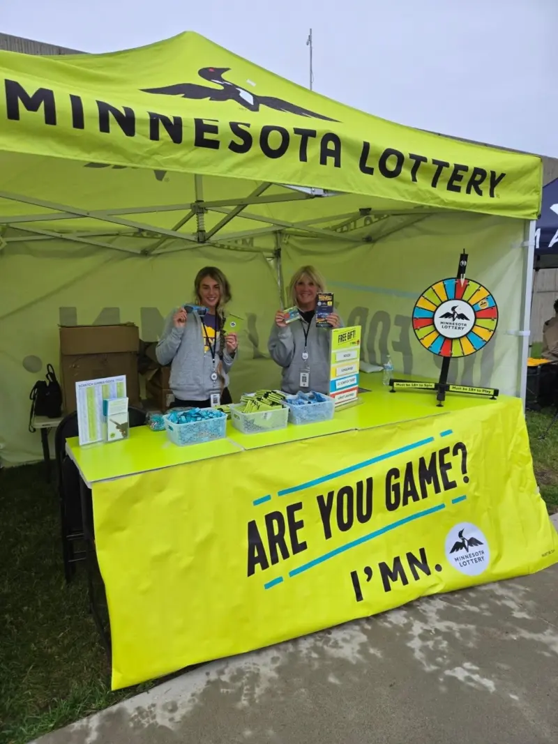 Two women hold lottery tickets under a yellow Minnesota Lottery tent with a prize wheel and “ARE YOU GAME? IMN” table display.