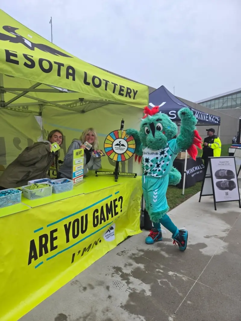 Two people and a teal mascot pose at a yellow Minnesota Lottery booth with a prize wheel. Booth reads "ARE YOU GAME?" Outdoors.