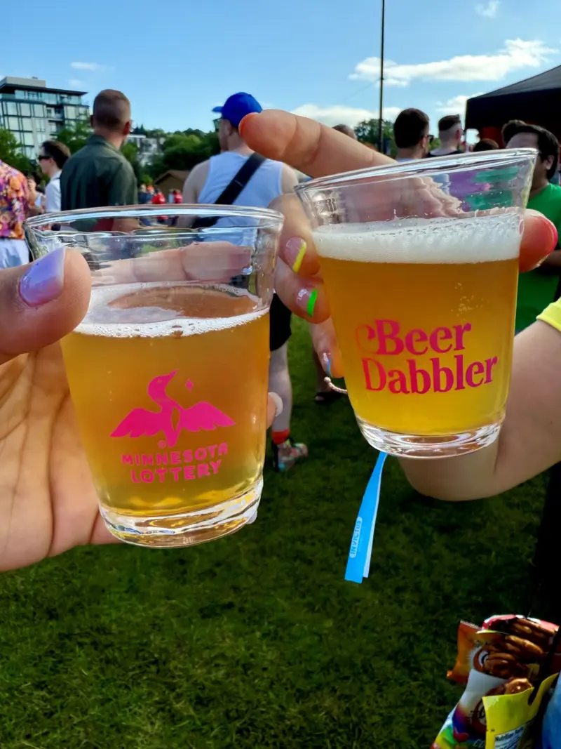 Two people toast with plastic beer cups—one marked Minnesota Lottery with a loon logo, the other Beer Dabbler—at an outdoor event.