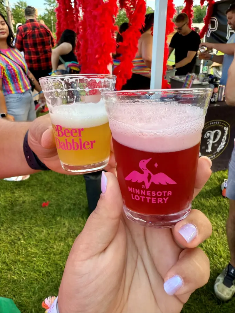 Two people toast with beer glasses labeled Beer Dabbler and Minnesota Lottery at an outdoor event with tents and crowds in the background.