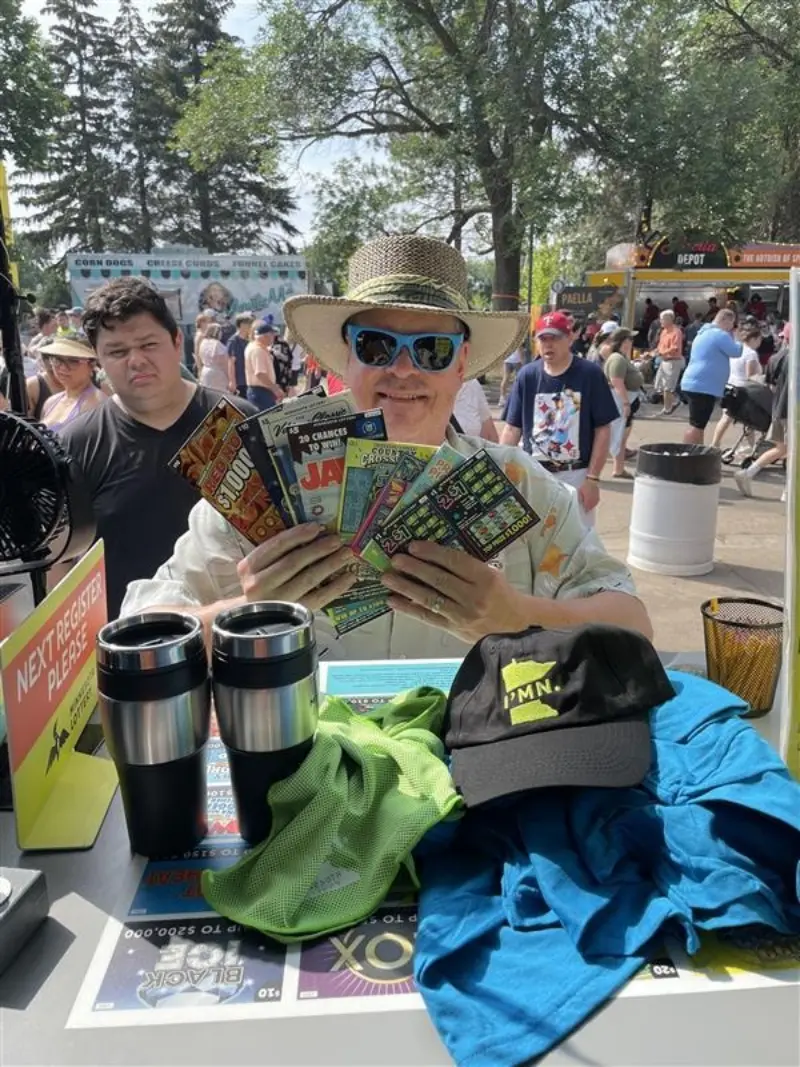 Person in sunglasses and straw hat holds colorful scratch-off tickets at outdoor event with crowds, vendor booths, and table items visible.