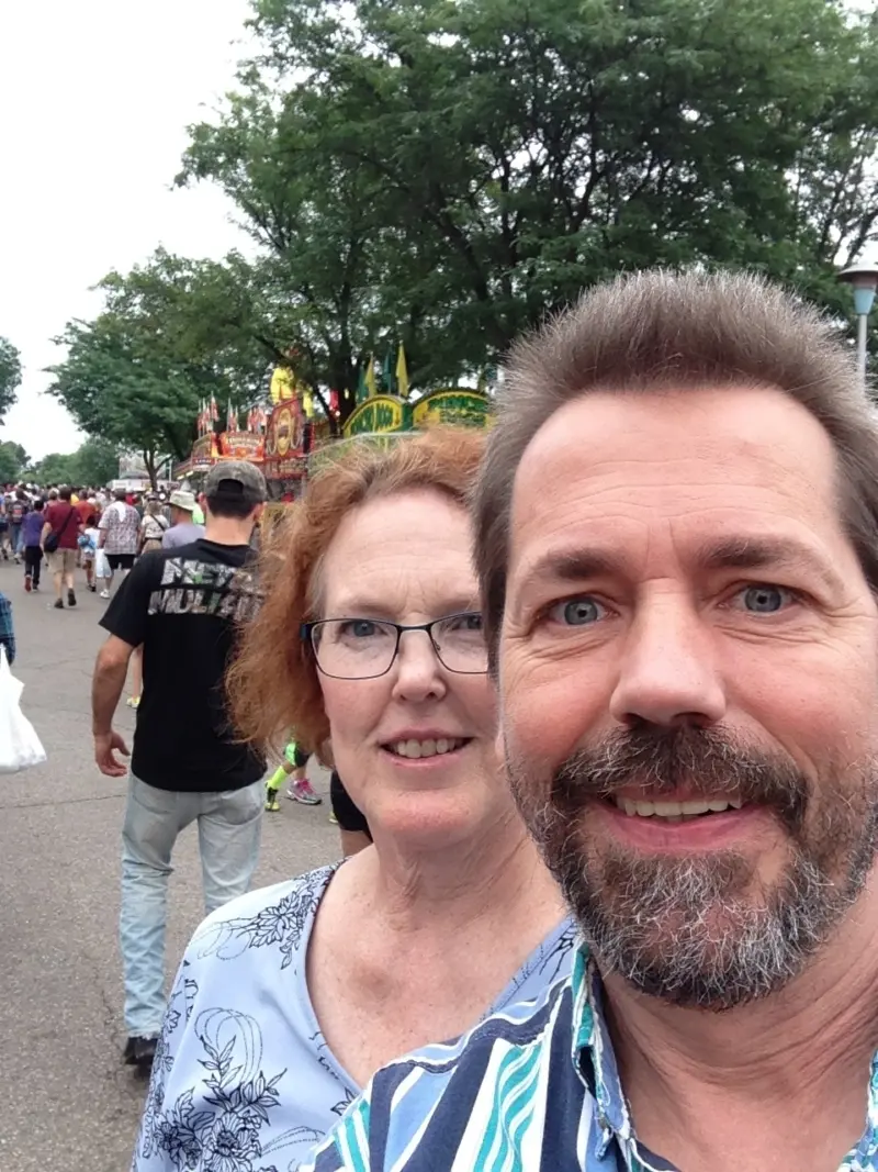 Smiling man and woman take a selfie at an outdoor fair, with food stands, trees, and people walking along a path in the background.