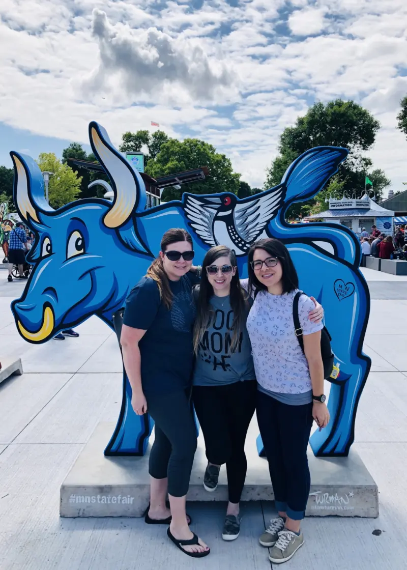 Three women smile in front of a blue ox statue with a bird on its back at an outdoor fair, trees and people visible under a partly cloudy sky.