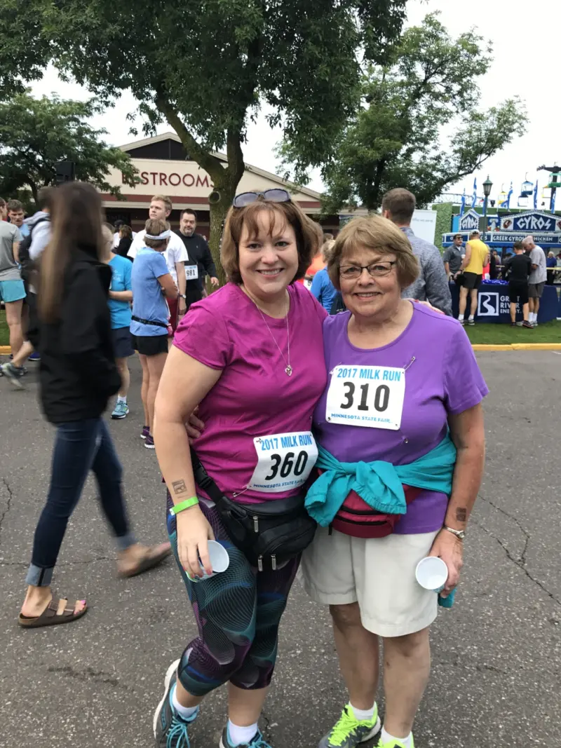 Two women in athletic wear with race numbers 360 and 310 smile, holding cups at an outdoor event with other participants and trees behind them.