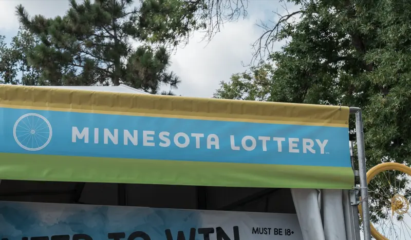Green and blue Minnesota Lottery sign with a wheel graphic at an outdoor event, trees and cloudy sky in the background.